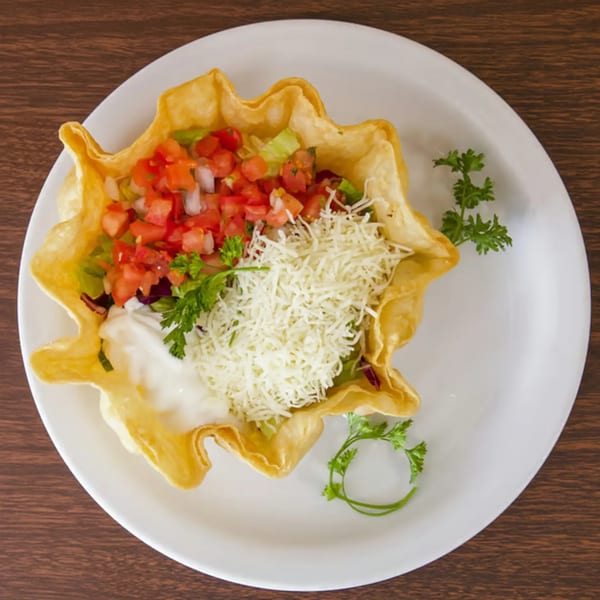 A taco salad in a crispy tortilla bowl, topped with diced tomatoes, shredded cheese, and sour cream.